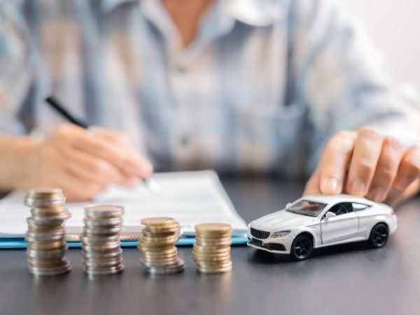 A model car placed beside stacks of coins on a desk, with a person writing in the background.