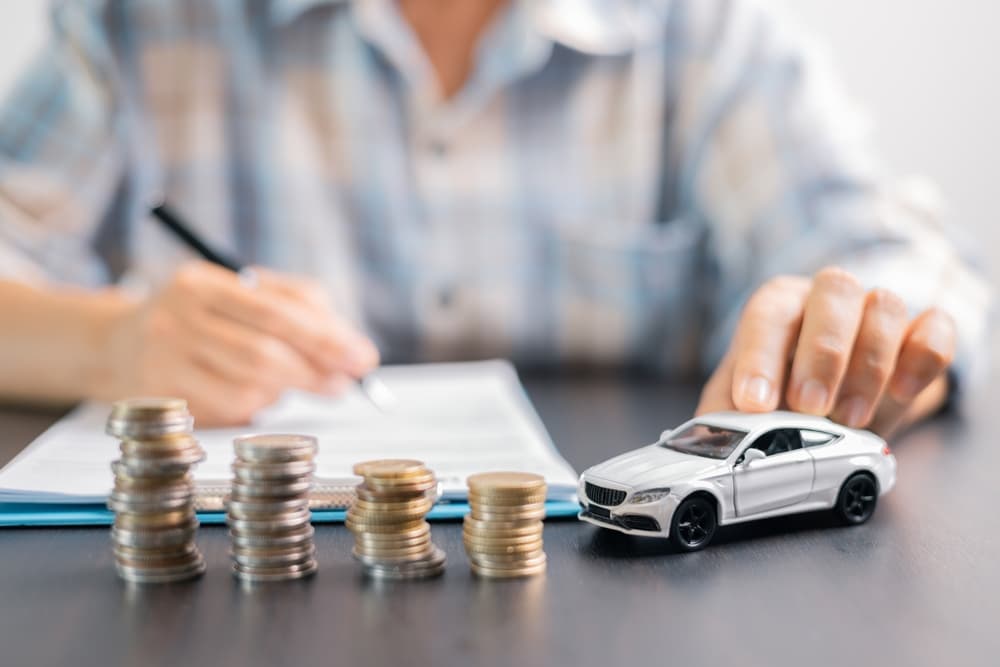 A model car placed beside stacks of coins on a desk, with a person writing in the background.